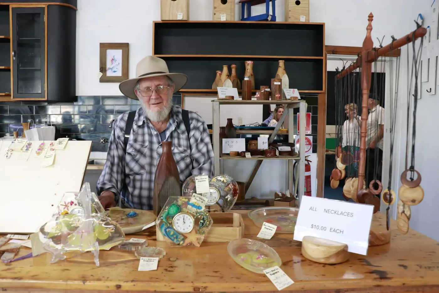 Peter Slaven from Eugowra Men\\u2019s Shed holding a vase made from a timber verandah post, surrounded by beautiful handmade items available at the store.